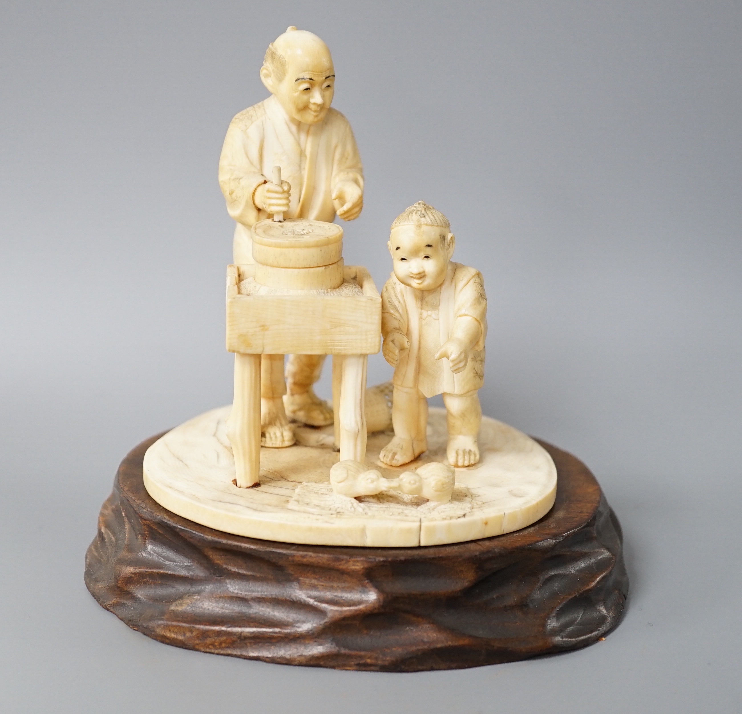 A Japanese ivory okimono of man and boy at a grindstone, Meiji period, signed, wood stand. 15cm total height